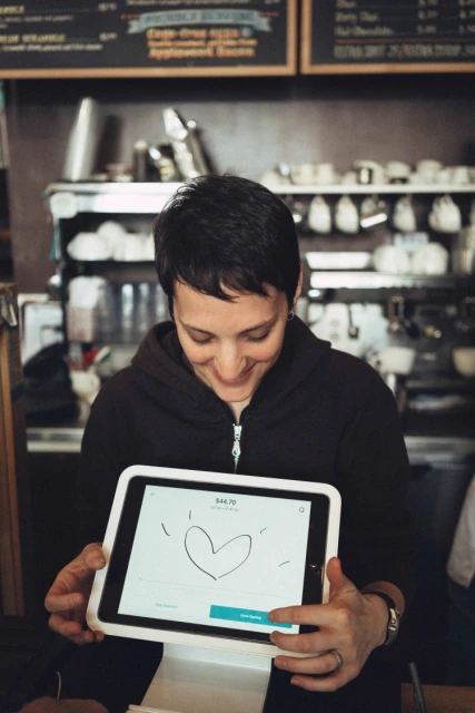Adult female employee holds a tablet with a heart drawing, smiling in an indoor setting, with cafe items in the background.