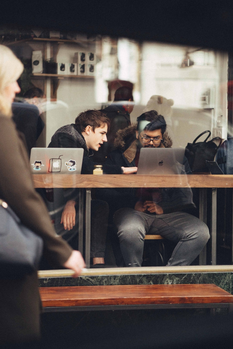 Two people work on laptops at a wooden table inside a cafe with shelving; a woman walks past the window.