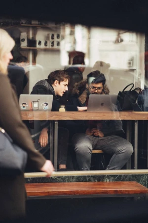 Two people work on laptops at a wooden table inside a cafe with shelving; a woman walks past the window.