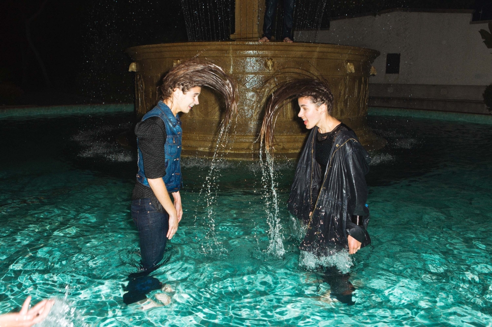 Two young adults standing in a fountain at night, flipping their wet hair in unison with a water feature in the background.