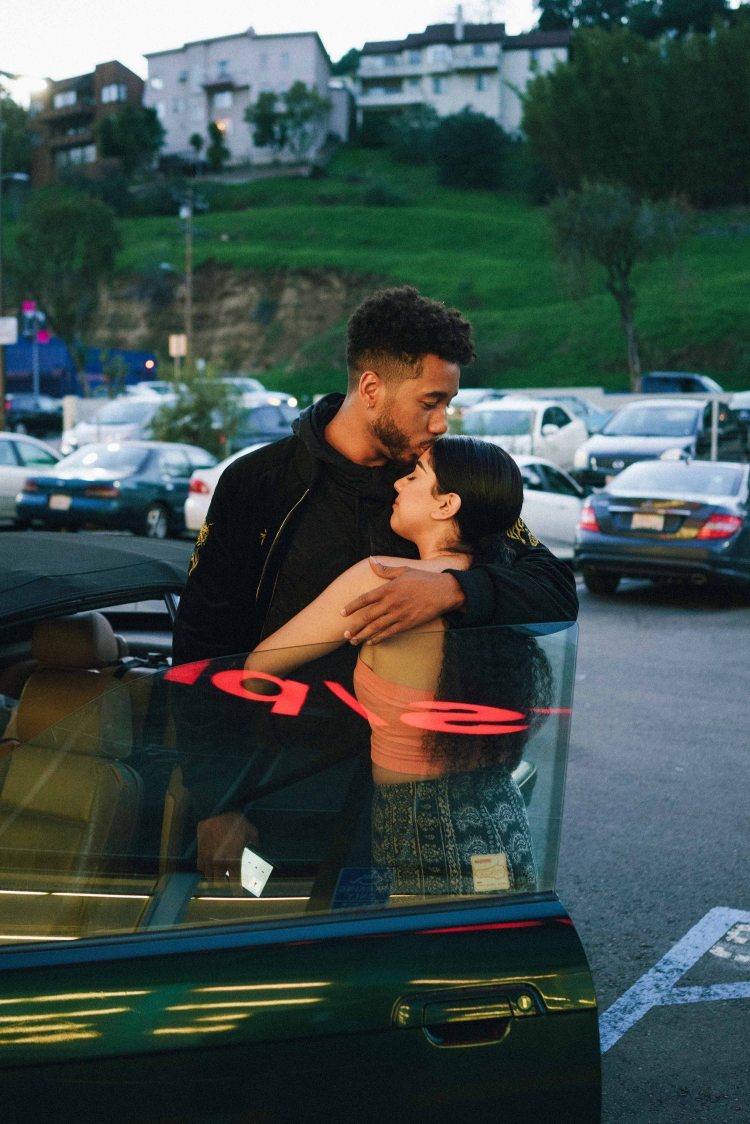 Intimate embrace between a man and woman in front of a car in a parking lot at dusk, with a hillside residential area visible.