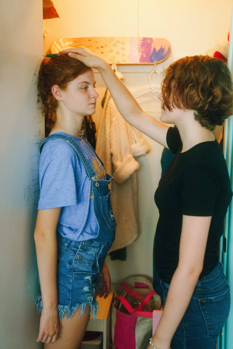 Two girls measure height on wall in closet. One holds pencil to mark level as the other stands against the wall, wearing denim overalls.