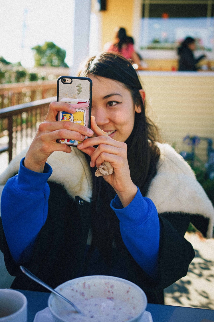 A woman holds a smartphone in front of her face to take a photo, sitting outside on a sunny day, with a bowl and spoon on the table.