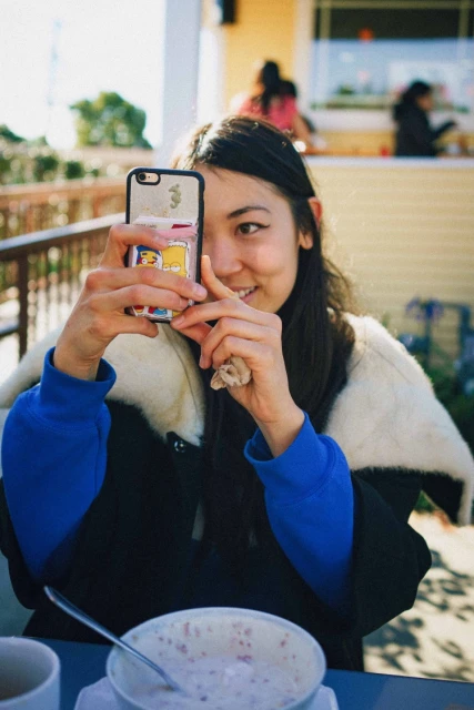 A woman holds a smartphone in front of her face to take a photo, sitting outside on a sunny day, with a bowl and spoon on the table.