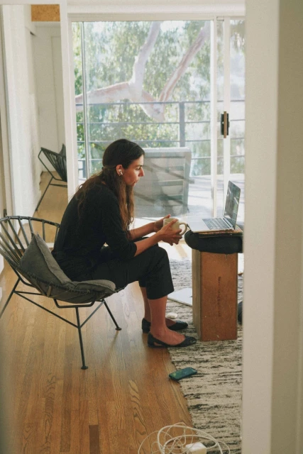 Woman sitting in a modern chair, working on a laptop in a bright indoor space with a view through open glass doors to the outside.