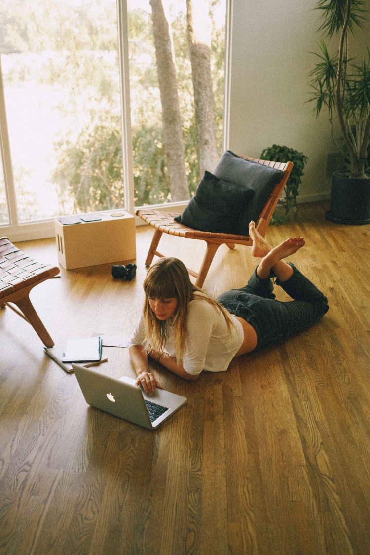 Woman lying on floor using laptop in bright airy room with large windows and hardwood floor, wooden chairs, and potted plants.