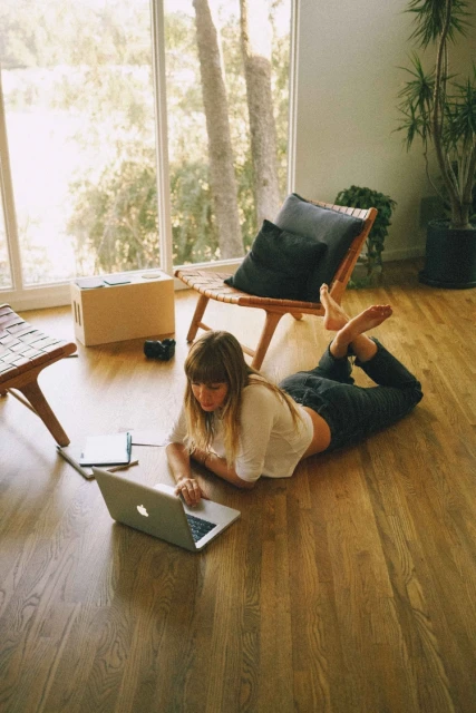 Woman lying on floor using laptop in bright airy room with large windows and hardwood floor, wooden chairs, and potted plants.