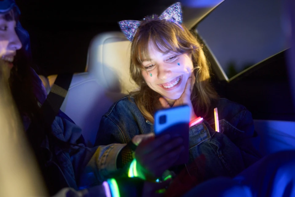 Two smiling young women inside car at night, looking at smartphone screen, wearing party accessories and glowing bracelets in blue light.
