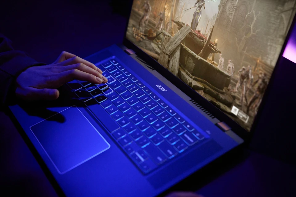 Close-up of person's hands typing on an Acer laptop with blue backlit keyboard; gaming displayed on the screen; dark indoor environment.