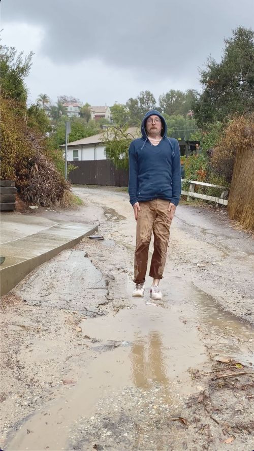Man wearing a blue hooded sweatshirt and brown cargo pants standing on a wet dirt road under an overcast sky