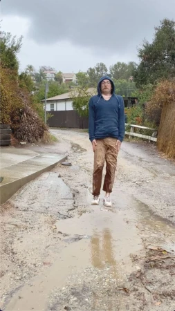 Man wearing a blue hooded sweatshirt and brown cargo pants standing on a wet dirt road under an overcast sky