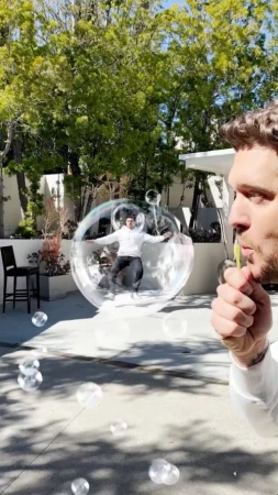 A man blows bubbles outside on a sunny day, with another man inside a large bubble in the background. Trees and a house are visible.