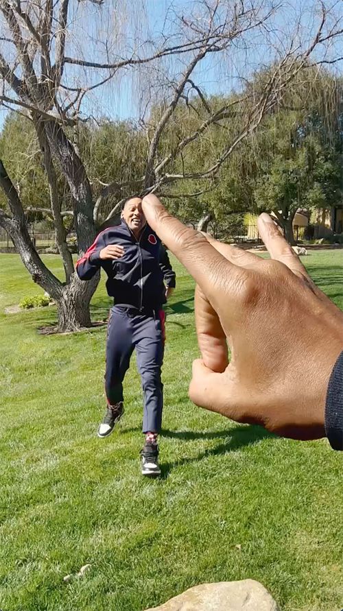 Outdoor shot of a man in a tracksuit running on grass with a large disembodied hand in the foreground on a sunny day.