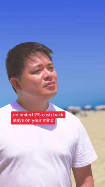 Man wearing a white t-shirt stands on a beach with blue sky and blurred beachgoers in the background, under bright sunlight.