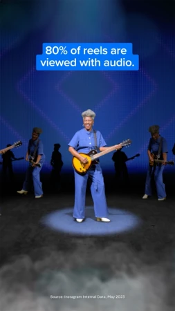 Adult woman playing electric guitar on stage surrounded by bandmates in blue jumpsuits with stage lighting and blue background.