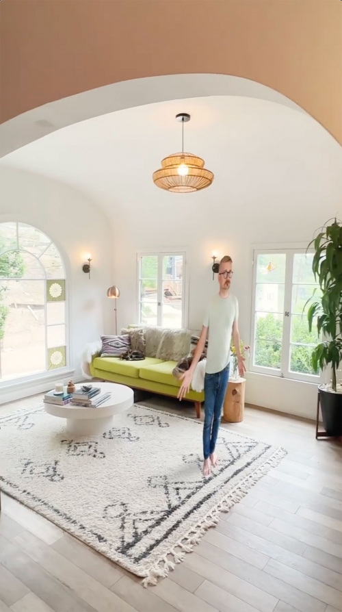 Adult male stands in a bright living room with a patterned rug, green sofa, coffee table, and a view through the window to the outdoors.