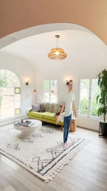 Adult male stands in a bright living room with a patterned rug, green sofa, coffee table, and a view through the window to the outdoors.