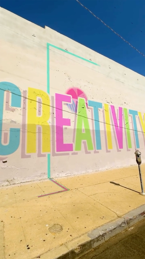 Outdoor mural featuring the colorful word 'CREATIVITY' in yellow, pink, and blue with a light blue border on a building wall.