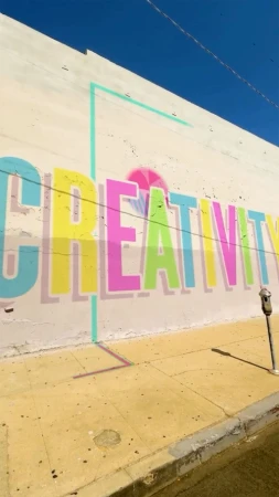 Outdoor mural featuring the colorful word 'CREATIVITY' in yellow, pink, and blue with a light blue border on a building wall.