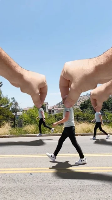 Optical illusion with large hands picking up tiny people walking on a road against a blue sky, creating a whimsical, surreal scene.