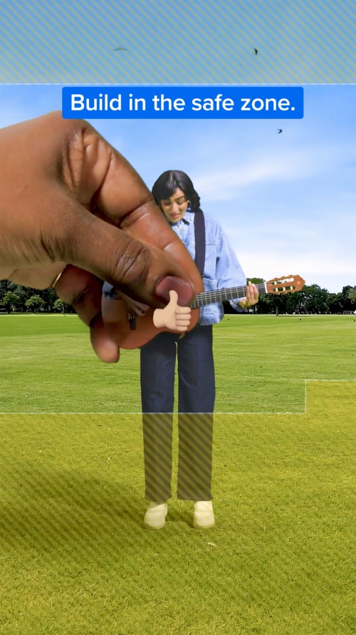 Woman plays guitar in sunny field with disembodied hand holding thumbs-up button, sky with clouds in the background.