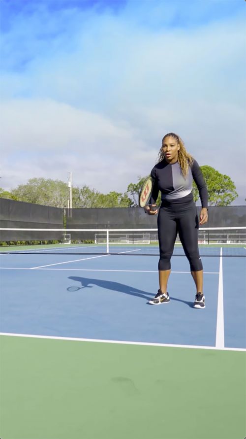 Serena Williams playing tennis outdoors on a blue and green court with a tennis racket in daylight with blue sky and clouds above.