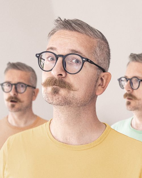 Studio shot of three adult men with moustaches and glasses, wearing yellow and green t-shirts against a plain light-toned background.