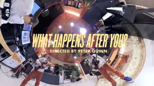 Music video still featuring two men playing electric guitars in an indoor setting with musical equipment and text overlay.
