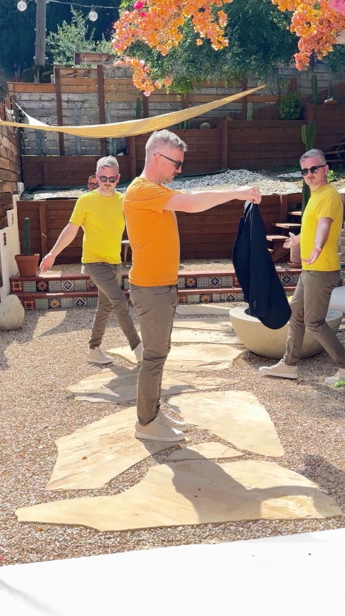 Adult male clones strike a pose on a stone-paved patio. Each man wears a differently colored shirt and dark blazer slung over the shoulder.