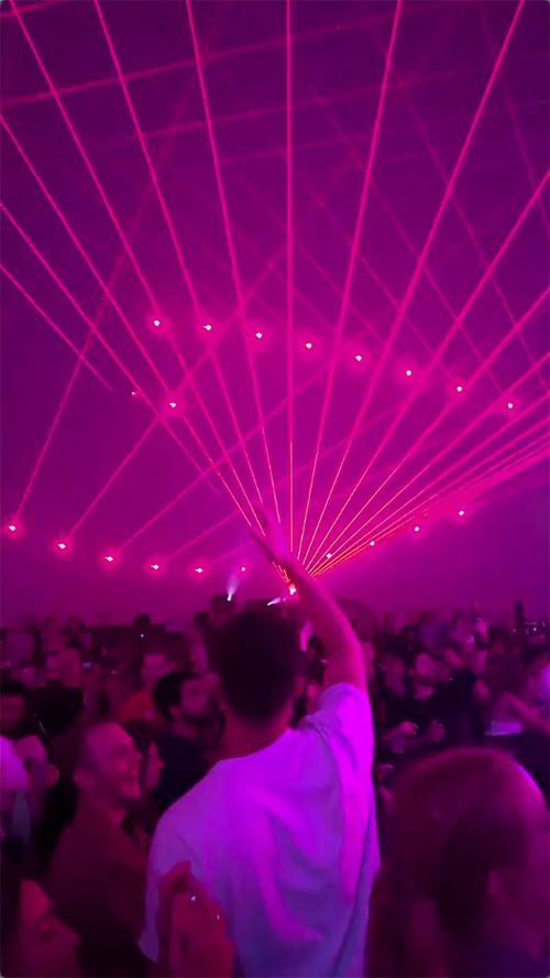 Large crowd of people at a concert with pink lasers, viewed from the back of a man raising his hand, capturing the vibrant energy of the event.