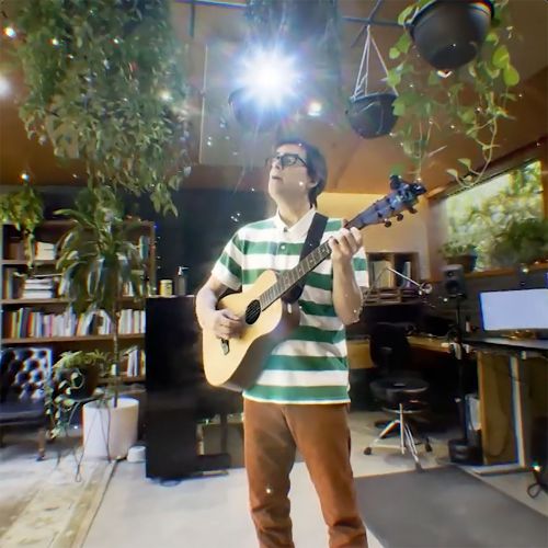 Man plays acoustic guitar in a sunlit room filled with plants, bookshelves, and music equipment, creating a harmonious, musical atmosphere.