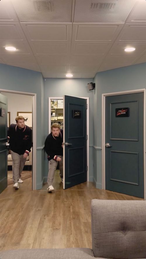 Two men emerge from separate doorways in an indoor hallway with neutral-toned walls and wooden floors, suggesting a candid or humorous moment.