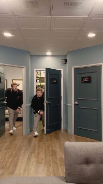 Two men emerge from separate doorways in an indoor hallway with neutral-toned walls and wooden floors, suggesting a candid or humorous moment.