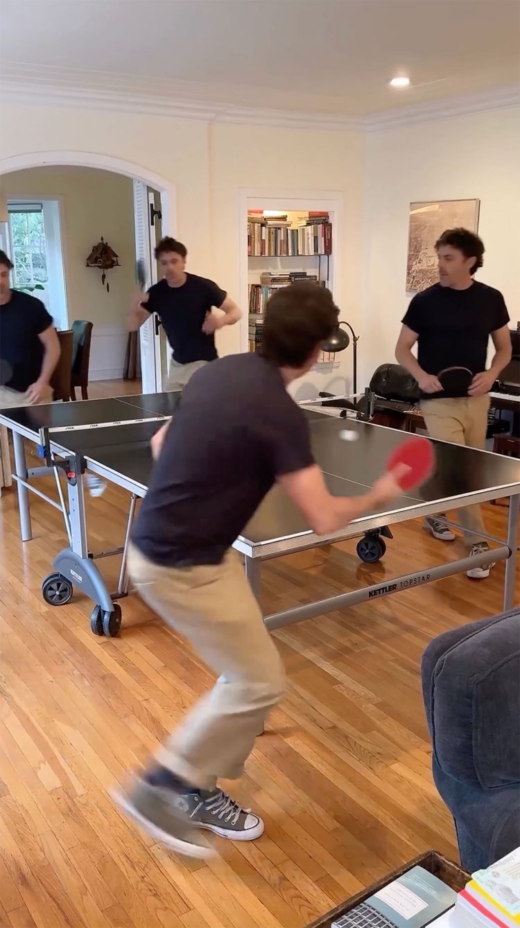 Young man playing table tennis indoors in a well-lit room with wooden floors and cream colored walls, multiple players visible in background.