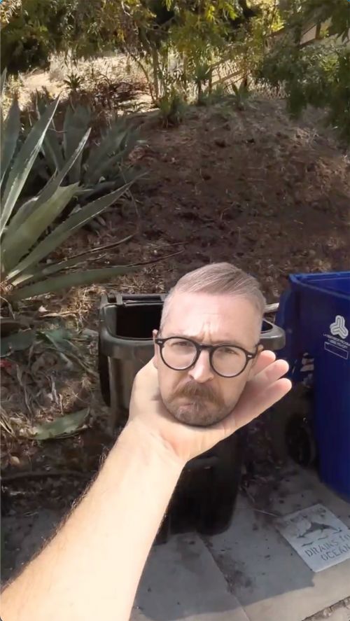 A person holds a man's head in their palm in an outdoor setting with lush greenery, waste bins, and an abstract visual effect.
