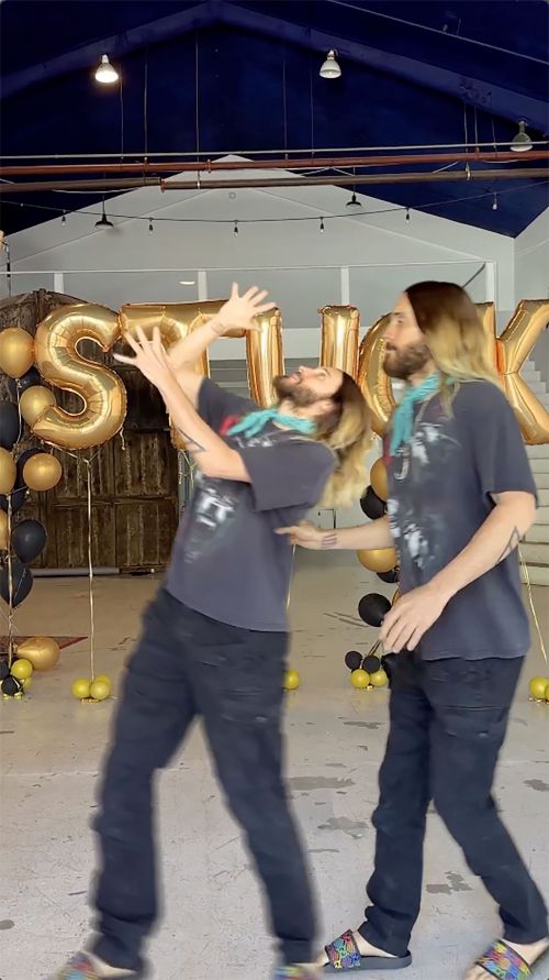 Two adult males in casual clothing standing indoors near gold balloon letters. The two men wear t-shirts and jeans in an indoor setting.