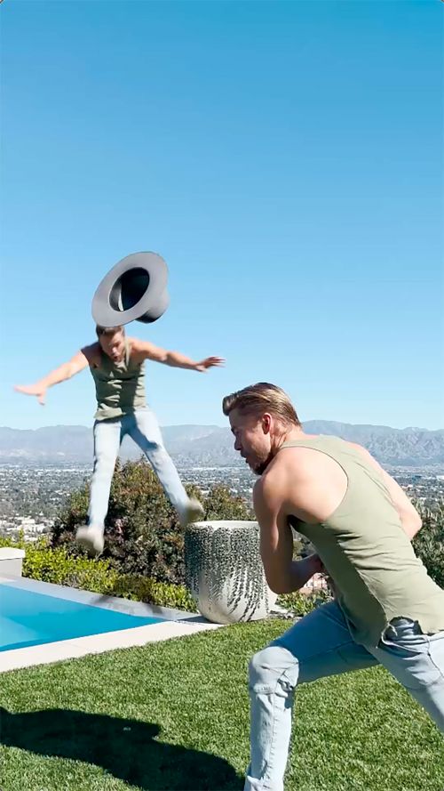 Two adult men in casual wear jumping and playing outside on a lawn, under a clear blue sky, with mountains in the distance.