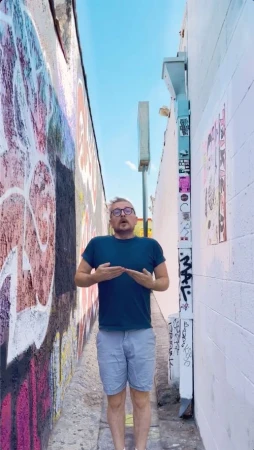 Adult male standing in alleyway with blue sky wearing glasses and shorts, flanked by graffiti and white wall in vertical shot.
