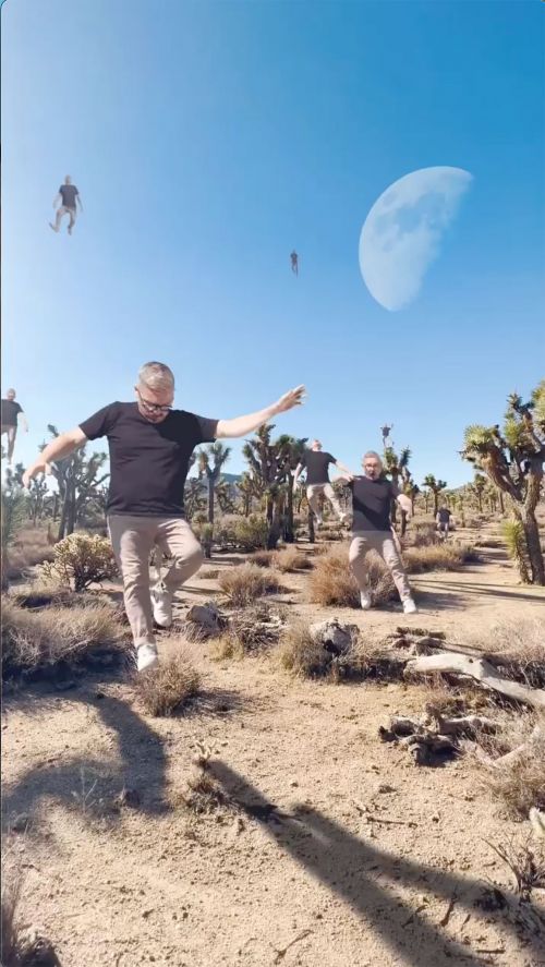 Man in black shirt and khaki pants balances along a desert path with other transparent clones of himself. Moon in the sky.