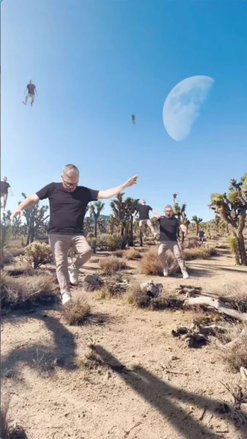 Man in black shirt and khaki pants balances along a desert path with other transparent clones of himself. Moon in the sky.