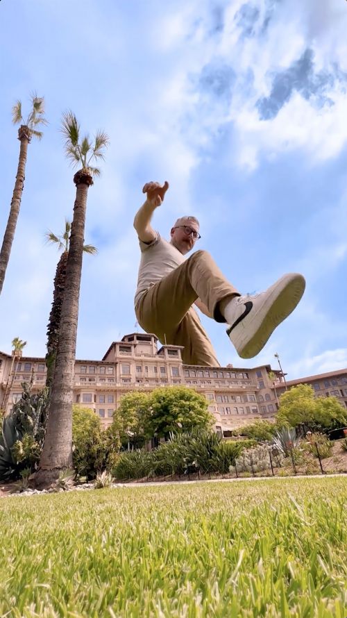 Man posing with architectural building. Person jumping with arms out. Green grass and palm trees surround hotel exterior on sunny day.