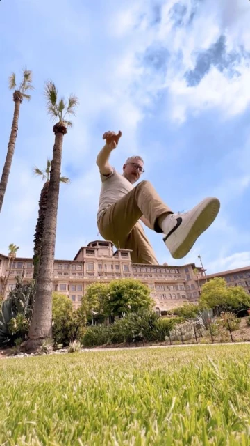 Man posing with architectural building. Person jumping with arms out. Green grass and palm trees surround hotel exterior on sunny day.