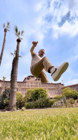 Man posing with architectural building. Person jumping with arms out. Green grass and palm trees surround hotel exterior on sunny day.