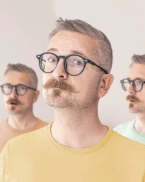 Close-up portrait of a man with round glasses, a mustache, and a yellow t-shirt, flanked by two similar figures.
