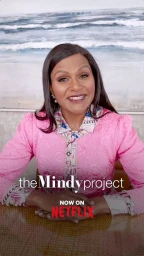 Mindy Kaling smiling in a pink patterned dress with a seascape painting in the background for The Mindy Project