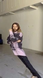 Jared Leto leaning dramatically at an impossible angle while wearing a pink and black graphic hoodie and dark trousers.