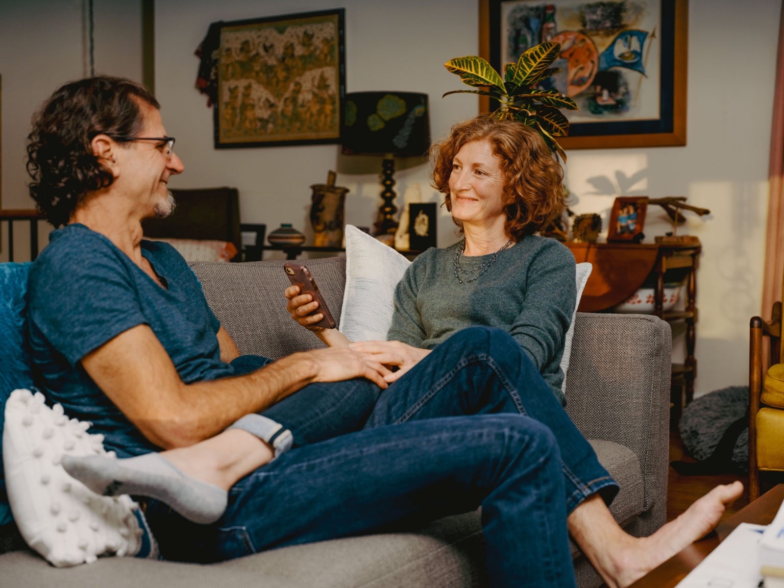 A relaxed couple sits on a gray sofa in a well-lit living room, casually dressed and enjoying each other's company at home.