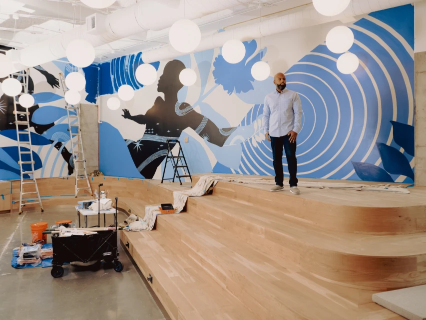 Man stands on wood platform in front of a mural of a woman with blue accents and geometric patterns, illuminated by globe lights.
