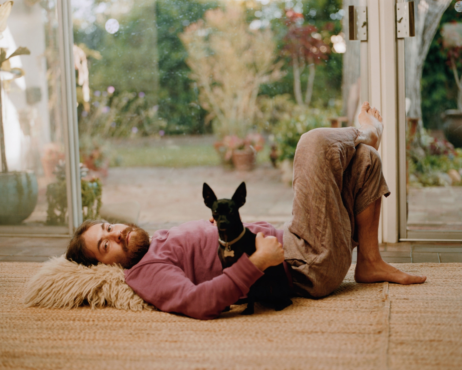 Bearded man in loungewear lying on a shag pillow with a black dog in an indoor sunroom, viewed through a wide angle lens with soft lighting.