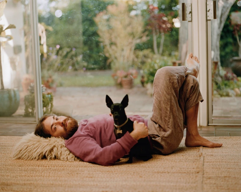 Bearded man in loungewear lying on a shag pillow with a black dog in an indoor sunroom, viewed through a wide angle lens with soft lighting.